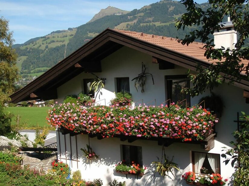 Alpines Wohnhaus mit blumengeschmücktem Holzbalkon vor bewaldeten Bergen.