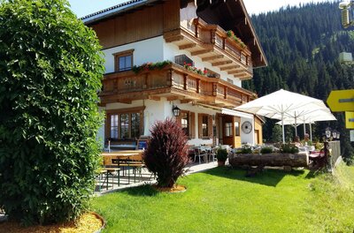 Alpines Gasthaus mit Holzfassade, Balkonen und einer Außenterrasse mit Sonnenschirmen vor bewaldetem Berg.