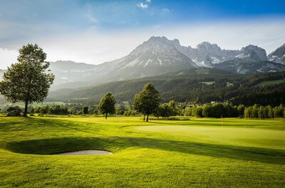 Eine malerische Golfanlage mit sattem Grün und sanften Hügeln. Im Hintergrund erheben sich majestätische Berge unter einem klaren Himmel.