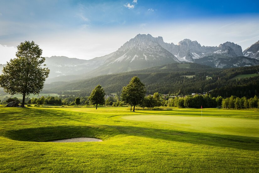 Ellmau_Golfschlaeger_Wilder Kaiser Eine malerische Golfanlage mit sattem Grün und sanften Hügeln. Im Hintergrund erheben sich majestätische Berge unter einem klaren Himmel.