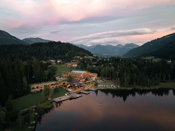 Luftaufnahme eines Hotels am Seeufer mit Bergen im Hintergrund bei Dämmerung.