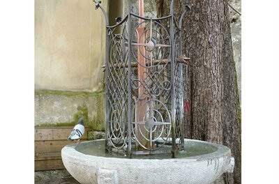 Ein dekorativer Brunnen aus Stein mit einem kunstvollen Gitter. Neben dem Brunnen sitzt eine Taube.