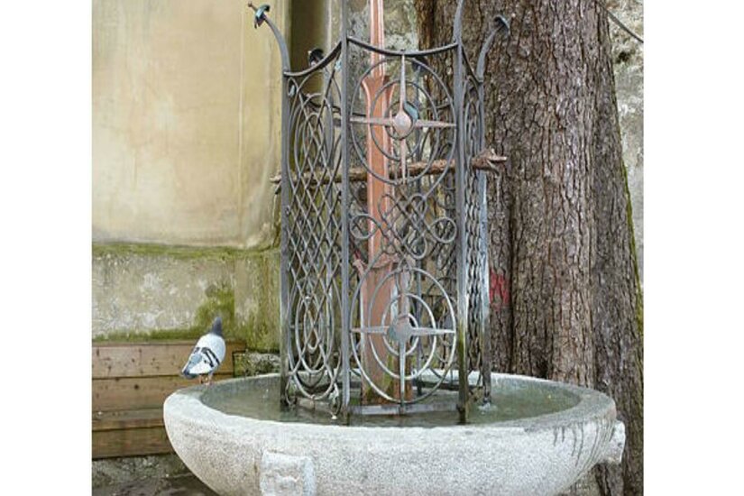 Ein dekorativer Brunnen aus Stein mit einem kunstvollen Gitter. Neben dem Brunnen sitzt eine Taube.