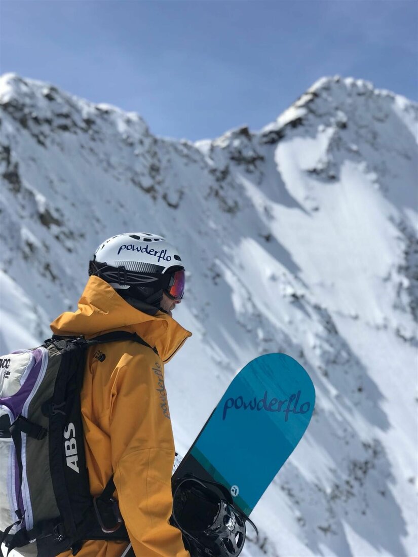 Snowboarder mit gelber Jacke, Helm und Rucksack blickt auf einen verschneiten Berggipfel.