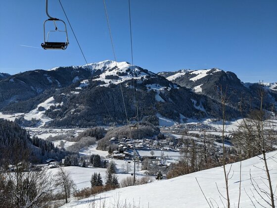 Sessellift bei Oberaigen Blick aus einem Sessellift auf ein verschneites Tal mit Dorf und bewaldeten Bergen unter blauem Himmel.