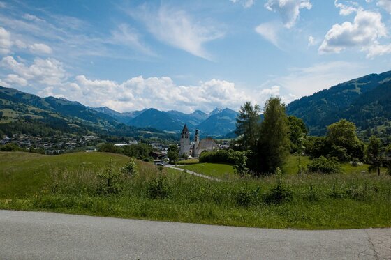 Blick über grüne Wiesen auf ein Dorf mit Kirchtürmen vor einer alpinen Bergkulisse unter blauem Himmel.