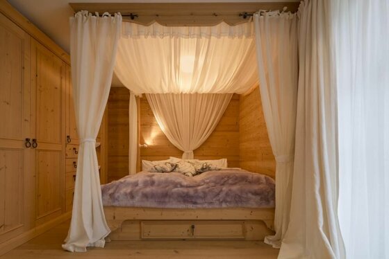 Bett Einliegerwohnung Chalet Bockberg Himmelbett aus hellem Holz mit weißen Vorhängen in einer holzgetäfelten Nische.