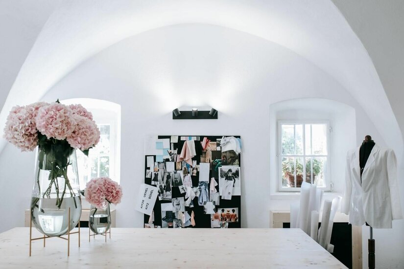 Atelier Simone Becke Heller Atelierraum mit Holztisch, rosa Hortensien in Glasvasen, Moodboard und Schneiderpuppe unter einem Gewölbe.