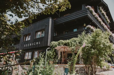 Außenansicht des Hotels Seebichl mit dunkler Holzfassade, Balkonen und einer begrünten Terrasse mit Sonnenschirm.