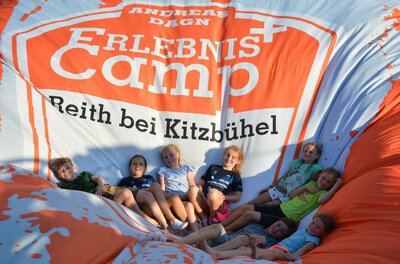 Kinder sitzen gemeinsam auf einem großen, orange-weißen Luftkissen mit der Aufschrift Erlebniscamp.