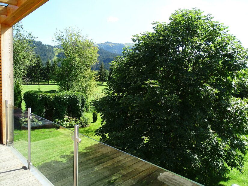 Blick von einem Holzbalkon mit Glasgeländer auf eine grüne Gartenanlage mit Bäumen und Bergen im Hintergrund.
