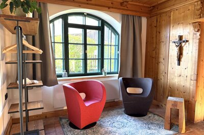 Ein gemütlicher Raum mit zwei modernen Sesseln in Rot und Schwarz. Ein großes Fenster sorgt für viel Licht und ein Holzregal ergänzt die stilvolle Einrichtung.