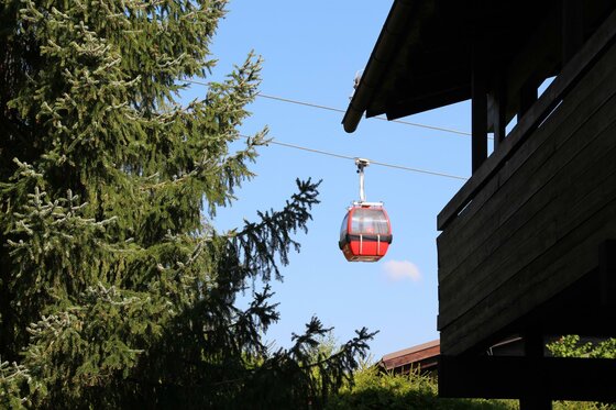 Seilbahn am Apartment Kitzkamm Rote Gondel einer Seilbahn schwebt vor blauem Himmel zwischen Nadelbäumen und einem Holzgebäude.