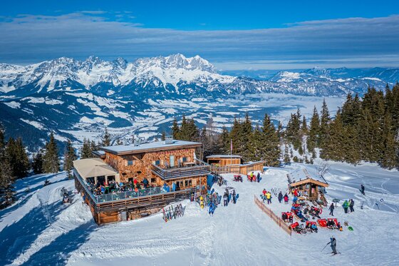 Berghütte aus Holz mit Sonnenterrasse in einem verschneiten Skigebiet vor markantem Gebirgspanorama.
