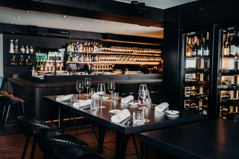Gedeckter dunkler Holztisch mit Weingläsern und Servietten vor einer Bar mit beleuchtetem Weinregal.
