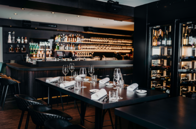 Gedeckter dunkler Holztisch mit Weingläsern und Servietten vor einer Bar mit beleuchtetem Weinregal.