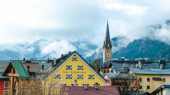 Stadtansicht Kitzbühel im Frühling Blick über Häuserdächer auf ein gelbes Hotelgebäude und einen Kirchturm vor bewölkten Bergen.