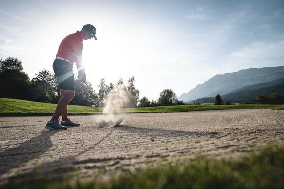 Ellmau_Golfer beim Abschlag_Wilder Kaiser Ein Golfer schlägt einen Ball aus dem Sandbunker. Im Hintergrund sind Bäume und Berge zu sehen.