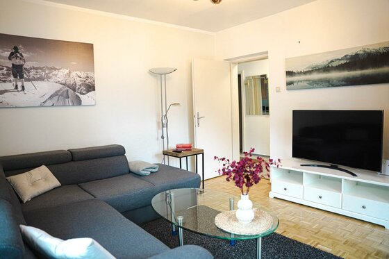 Ein gemütliches Wohnzimmer mit einer grauen Couch und einem Glastisch. An der Wand hängen Bilder von Berglandschaften, und ein Fernseher steht auf einem weißen TV-Tisch.