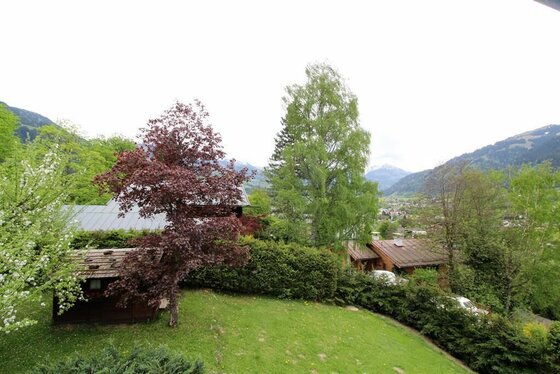 Gartenblick Lodge 12 Blick von oben auf einen Garten mit Rasenfläche, Bäumen und angrenzenden Häusern vor einer Bergkulisse.