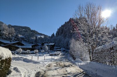 Winterliche Siedlung Oberaigen Verschneite Straße in einer alpinen Siedlung mit Holzhäusern und bewaldeten Bergen unter blauem Himmel.