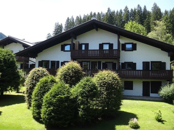 Zweistöckiges weißes Haus mit dunklen Holzbalkonen und grünen Sträuchern im Vordergrund.