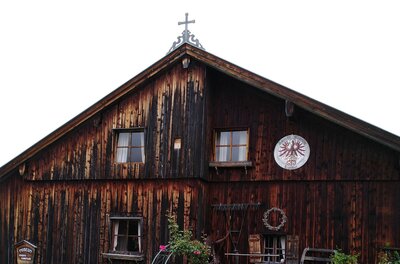 Bauernhausmuseum Vorderkünstler Eine rustikale Holzhütte mit einem Kreuz auf dem Dach. An den Wänden sind Fenster und ein Wappensymbol zu sehen.