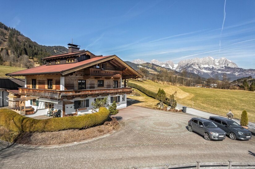Alpines Holzhaus mit Balkon vor einer Bergkette unter blauem Himmel.