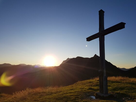 Ein hölzernes Gipfelkreuz auf einer Wiese vor einer Bergkulisse bei Sonnenuntergang.