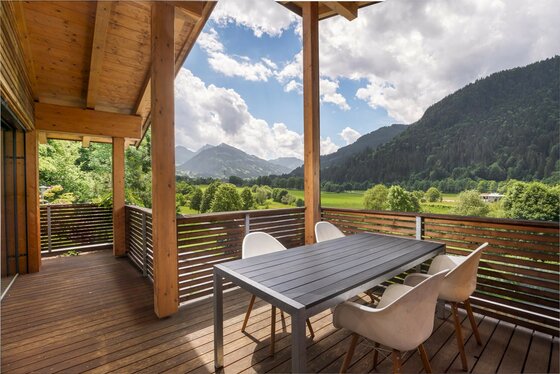 Holzbalkon mit Sitzgruppe und Blick auf bewaldete Berge und grüne Wiesen unter bewölktem Himmel.