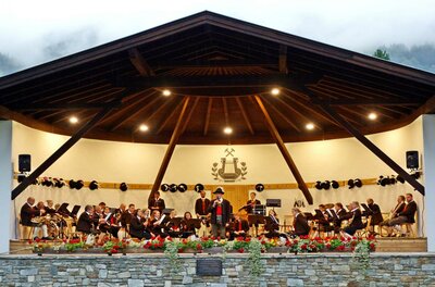 Pavillon Jochberg Eine Musikkapelle spielt auf einer Bühne in einer ansprechenden Umgebung. Im Hintergrund sind die Berge und ein bewölkter Himmel zu sehen.