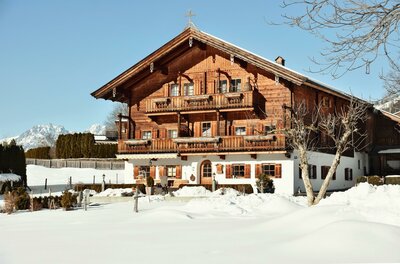 Ein traditionelles Holzhaus mit Balkonen in einer verschneiten Winterlandschaft vor Bergen.