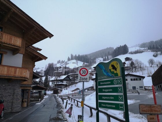 Verschneite Dorfstraße mit Hinweisschildern zur Bruggeralm und Bergbahnen vor bewaldetem Hang.
