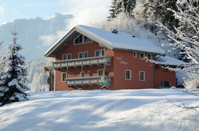 Ein rotes, schneebedecktes Gasthaus mit Holzbalkonen in einer winterlichen Berglandschaft.