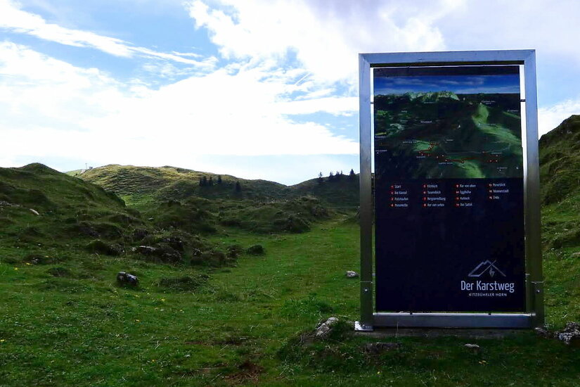 Infotafel am Karstweg Informationstafel zum Karstweg in einer grünen Hügellandschaft unter bewölktem Himmel.