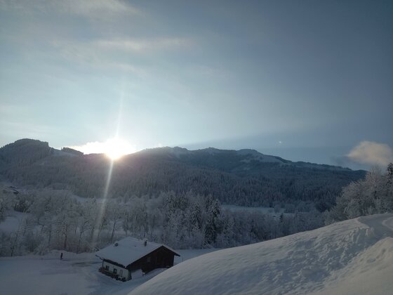 Eine winterliche Landschaft mit schneebedeckten Bergen und einem Sonnenaufgang. Im Vordergrund steht ein kleines, eingeschneites Haus.