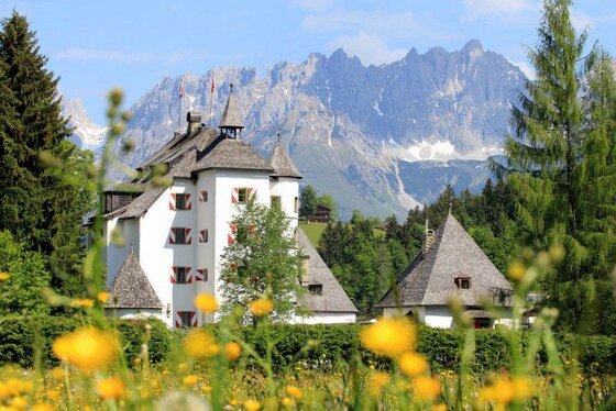 Sommeransicht Hotel Schloß Münichau Weißes Schloss mit rot-weiß gestreiften Fensterläden vor einem schroffen Gebirgsmassiv und einer Blumenwiese.