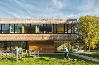 Modernes Holzgebäude mit Glasfront und davorliegendem Spielplatz mit balancierenden Kindern.