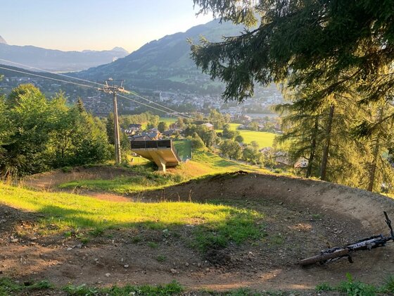Bike Trail am Hahnenkamm Blick auf eine Mountainbike-Strecke mit Steilkurven, im Hintergrund eine Seilbahnstütze und ein Tal mit Ortschaft.