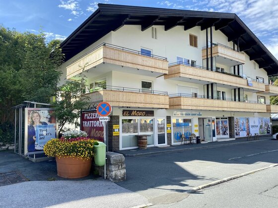Mehrstöckiges Gebäude mit Holz-Balkonen, einer Pizzeria im Erdgeschoss und einer Bushaltestelle davor.