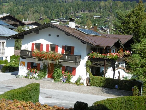 Alpines Wohnhaus mit weißen Wänden, roten Fensterläden und blumengeschmückten Holzbalkonen vor bewaldetem Hang.