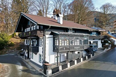 Traditionelles Alpenhaus mit dunkler Holzfassade, weißem Erdgeschoss und verzierten Balkonen vor bewaldetem Hang.