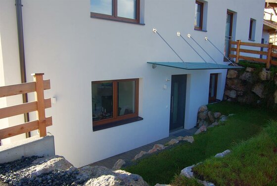 Hauseingang mit Glasvordach, weißer Fassade und Holzfenstern.