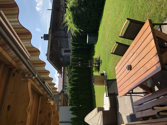 Terrasse Haus Hagleitner 2 Terrasse mit Holztisch, Stühlen und Markise vor einem Garten mit Rasenfläche und Hecke.