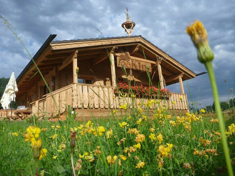 Café ecKing im Sommer Holzhütte mit Terrasse hinter einer Wiese mit gelben Blumen unter bewölktem Himmel.