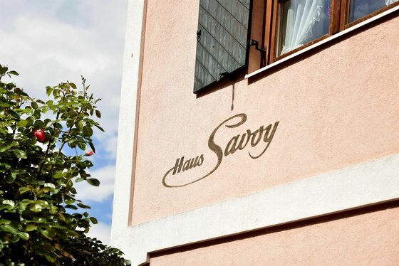 Rosa Hausfassade mit dem Schriftzug Haus Savoy, einem Fenster mit Fensterläden und einem Rosenstrauch im Vordergrund.