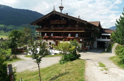 Großes, mehrstöckiges Holzhaus im alpinen Stil mit Blumenbalkonen vor bewaldeten Bergen.