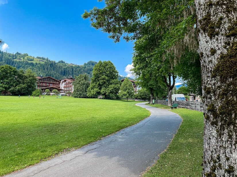 Stadtrunde Kitzbühel Asphaltierter Spazierweg führt an einer grünen Wiese und Bäumen vorbei in Richtung von Gebäuden im Hintergrund.