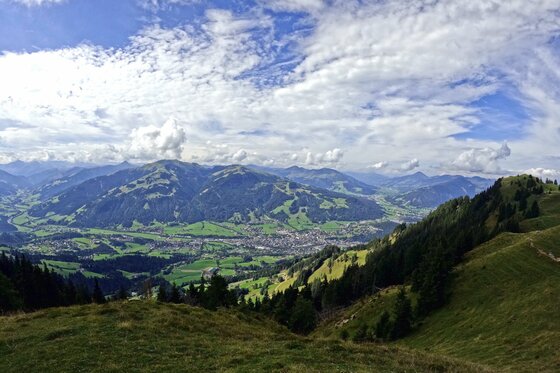 Aussicht vom Karstweg Panoramablick von einem grasbewachsenen Berghang auf ein bewohntes Tal und gegenüberliegende bewaldete Berge.