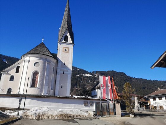Weiße Kirche mit spitzem Turm vor bewaldeten Bergen unter blauem Himmel.